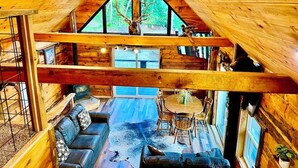A cozy cabin interior with a high ceiling, wooden beams, a fireplace, a dining area, and a large window with a deer head decoration.