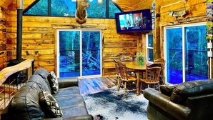 A log cabin interior with a wooden dining table, leather sofas, and a mounted deer head.