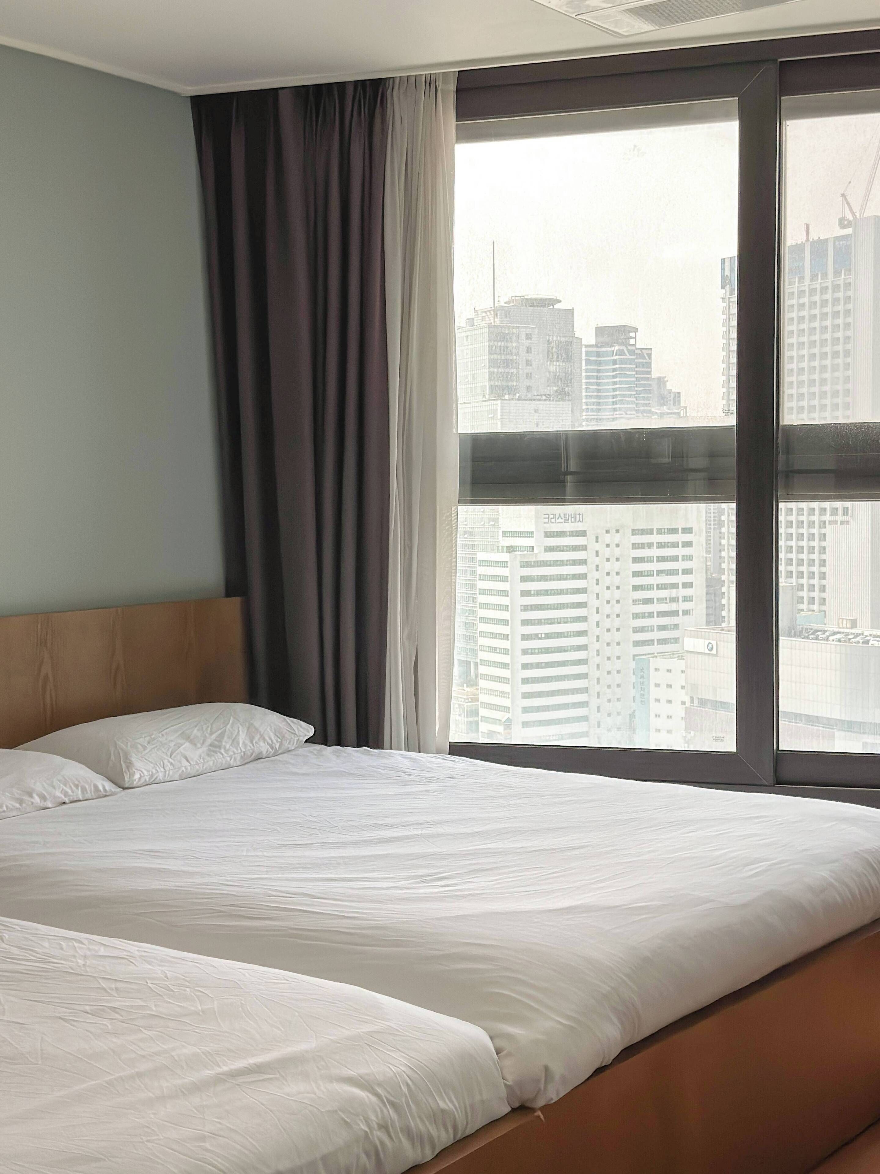 A hotel room with a large bed, a view of the cityscape through a window, and a curtain on the left side.