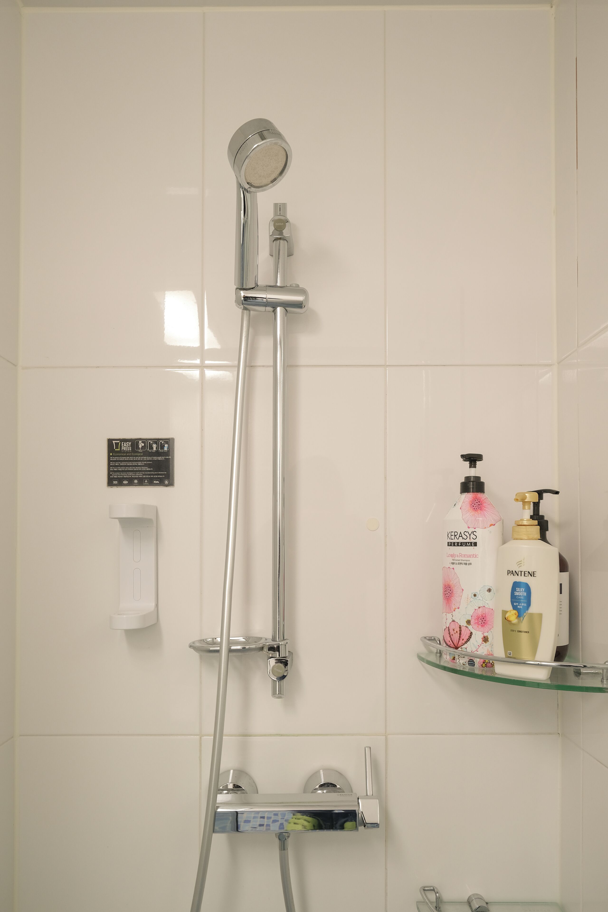 A shower with a handheld showerhead, a glass shelf with toiletries, and a white towel dispenser.