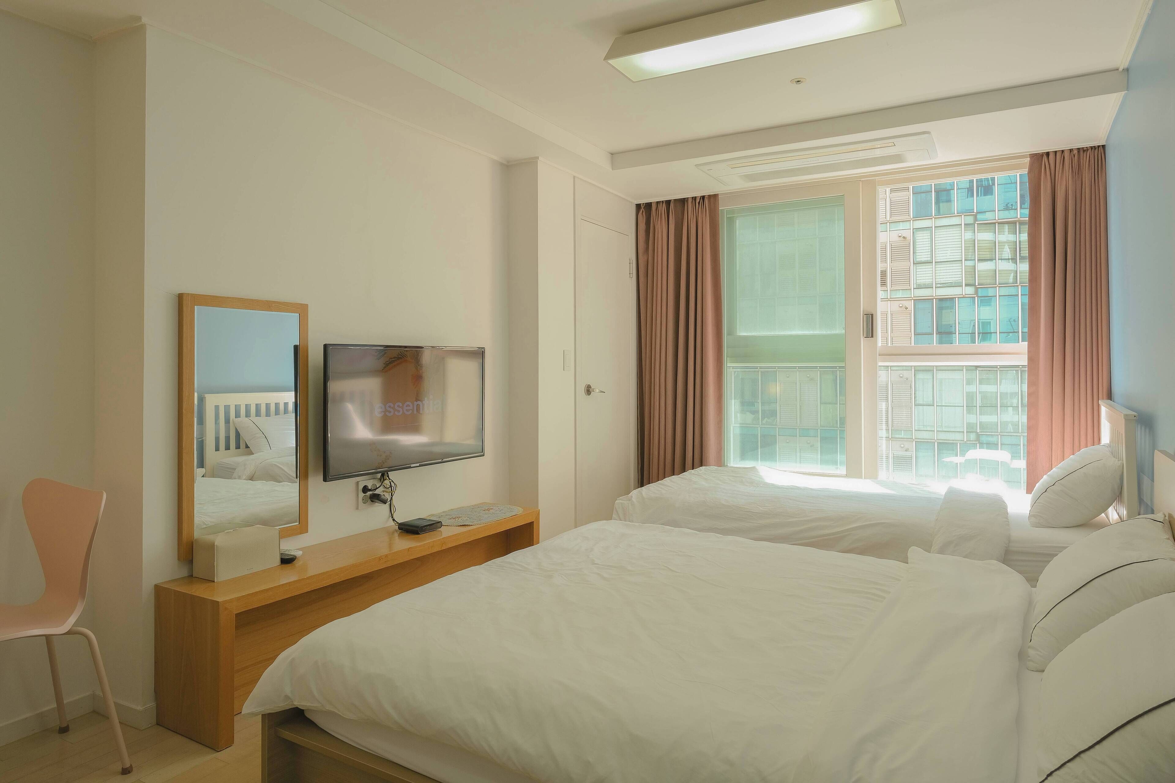 Une chambre d’hôtel avec deux lits, une télévision, un miroir, un bureau en bois et une fenêtre avec des rideaux.