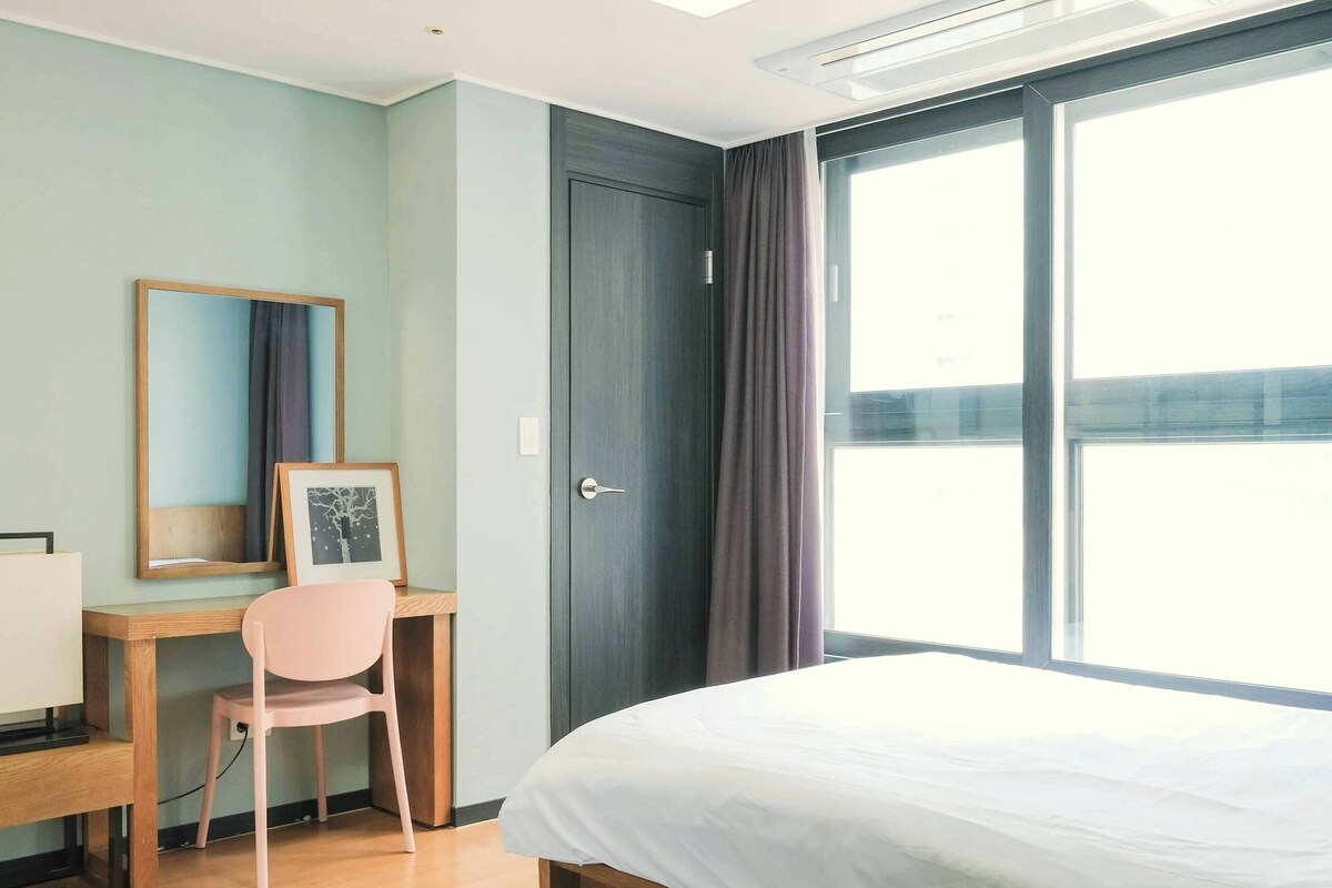 A modern hotel room with a large bed, a desk with a chair, a window with curtains, and a door.
