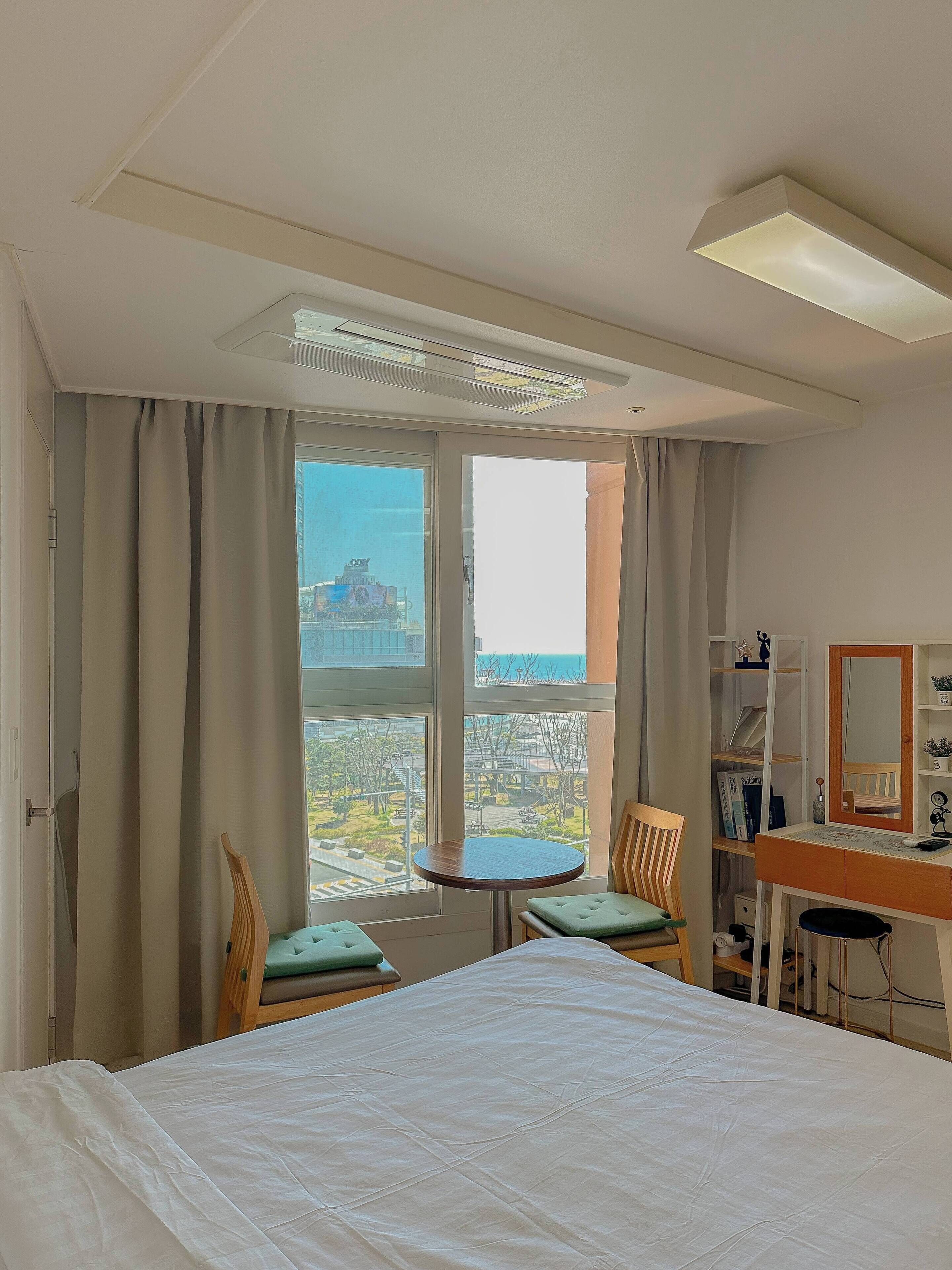 A hotel room with a bed, two chairs, a small table, a desk with a mirror, and a view of a cityscape through large windows.