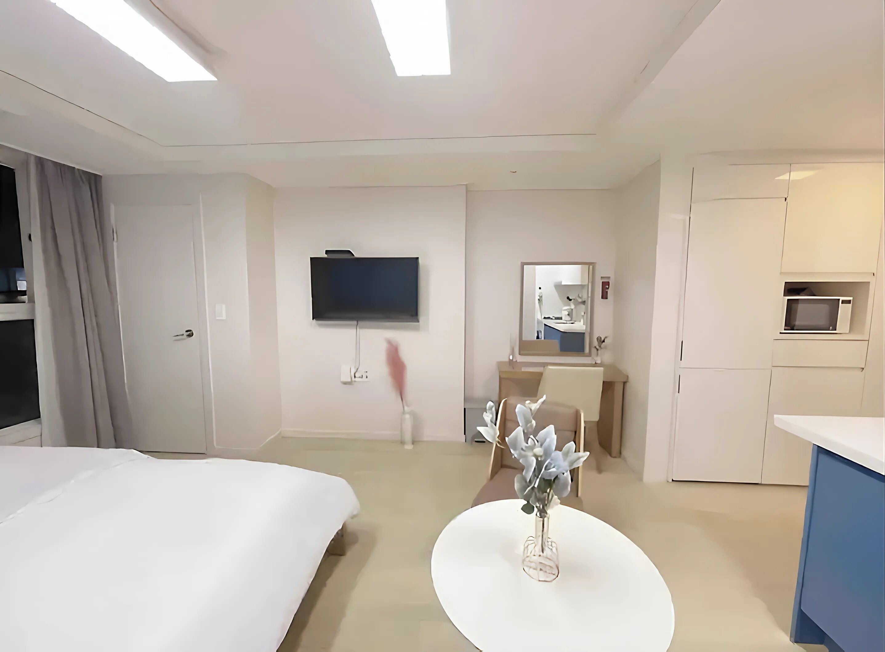 Habitación de hotel con cama, televisión montada en la pared, una mesita con un florero, una silla y una pequeña cocina.