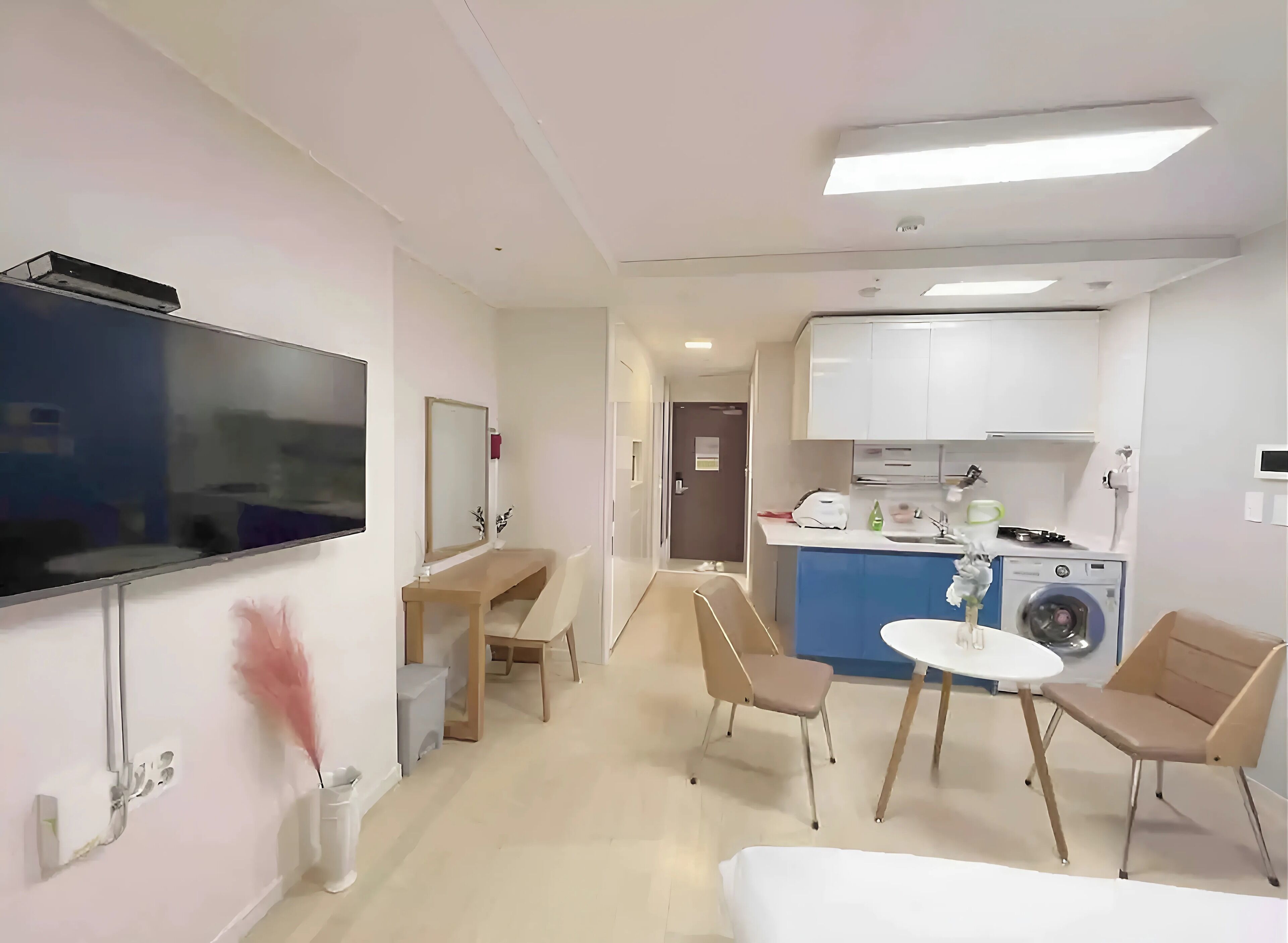Habitación de hotel pequeña con televisión, escritorio, silla, mesita, lavadora y kitchenette.