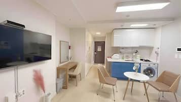 Habitación de hotel pequeña con televisión, escritorio, silla, mesita, lavadora y kitchenette.