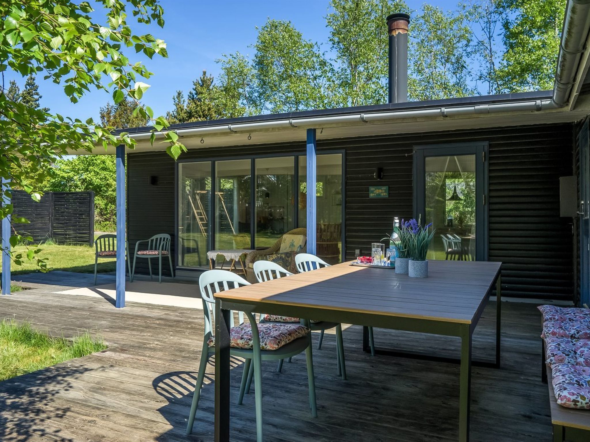 Une terrasse extérieure moderne, dotée d’une table et de chaises en bois, d’un espace salon couvert et offrant une vue sur le jardin arrière grâce à de grandes portes-fenêtres coulissantes.