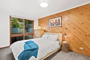 A bedroom with a bed, a bedside lamp, a framed picture on the wall, and a window with a view of greenery.