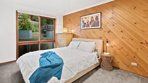 A bedroom with a bed, a bedside lamp, a framed picture on the wall, and a window with a view of greenery. - The Weekender With Spa Sleeps 11 (Ocean Grove)