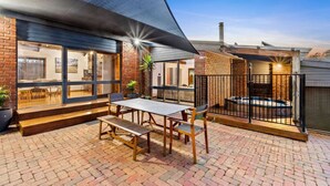 A patio area with a brick floor, a wooden table with benches, and a black metal fence. - The Weekender With Spa Sleeps 11 (Ocean Grove)