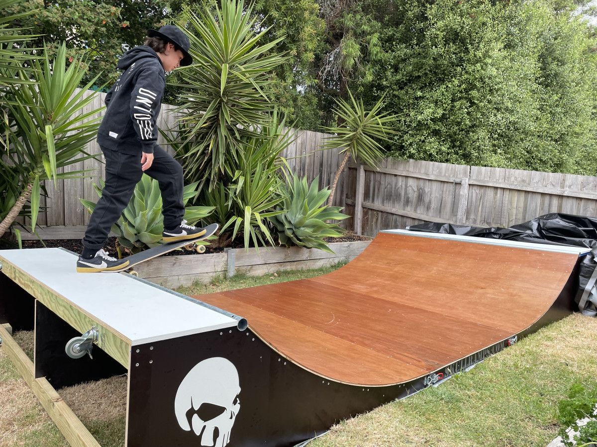 A person skateboarding on a ramp with a skull design, surrounded by a wooden fence and greenery.