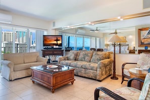 A spacious living room with a large flat-screen TV, comfortable sofas, a coffee table, and a view of the city skyline through large windows.