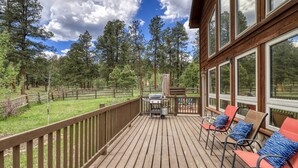 Une terrasse en bois avec des sièges d’extérieur et un barbecue, entourée d’une zone délimitée par une clôture et d’arbres.