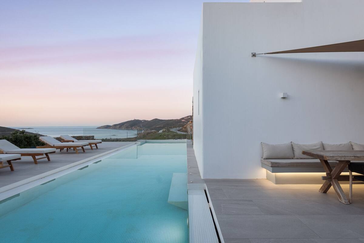 A modern infinity pool with lounge chairs and a wooden table, overlooking a scenic coastline.