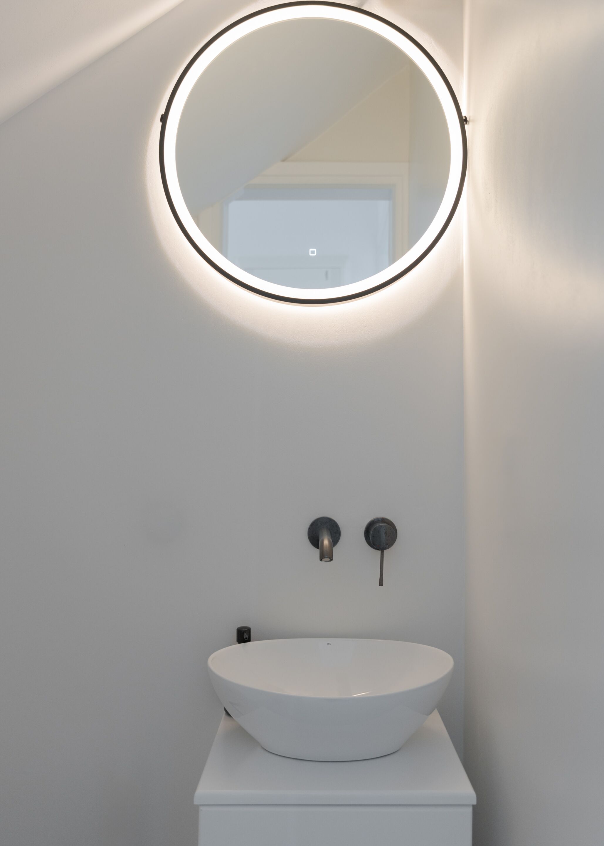 A bathroom with a white sink, a round mirror with a light, and two wall-mounted faucets.