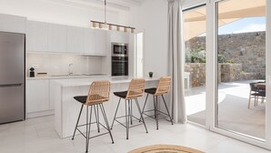 A modern kitchen with white cabinetry, a central island with bar stools, and a stone wall visible through the glass door. - Retreat Hill Luxury Villas Mykonos (Mykonos)