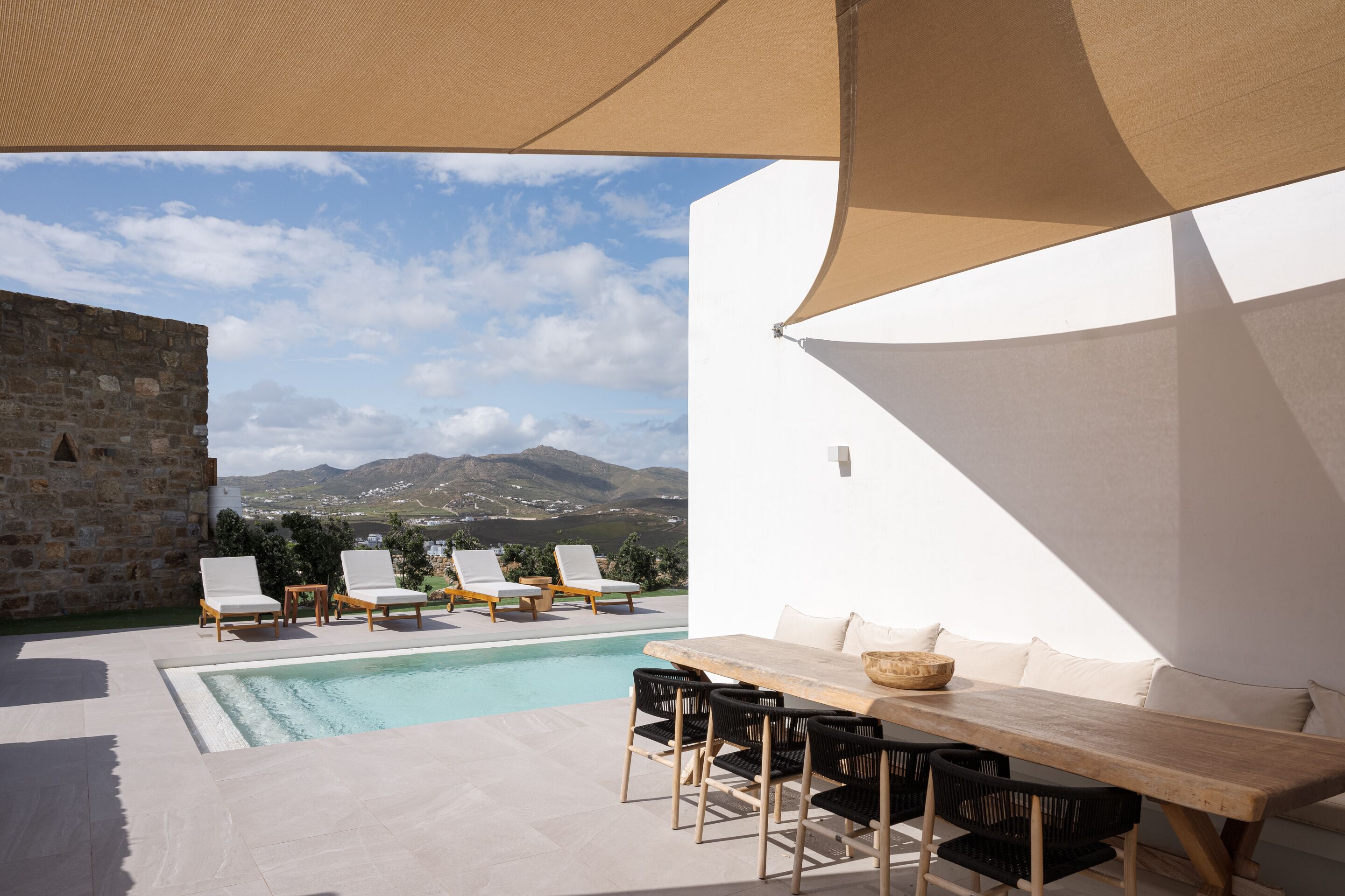 Zona junto a la piscina con sillones de descanso, una mesa de madera y vistas a montañas.