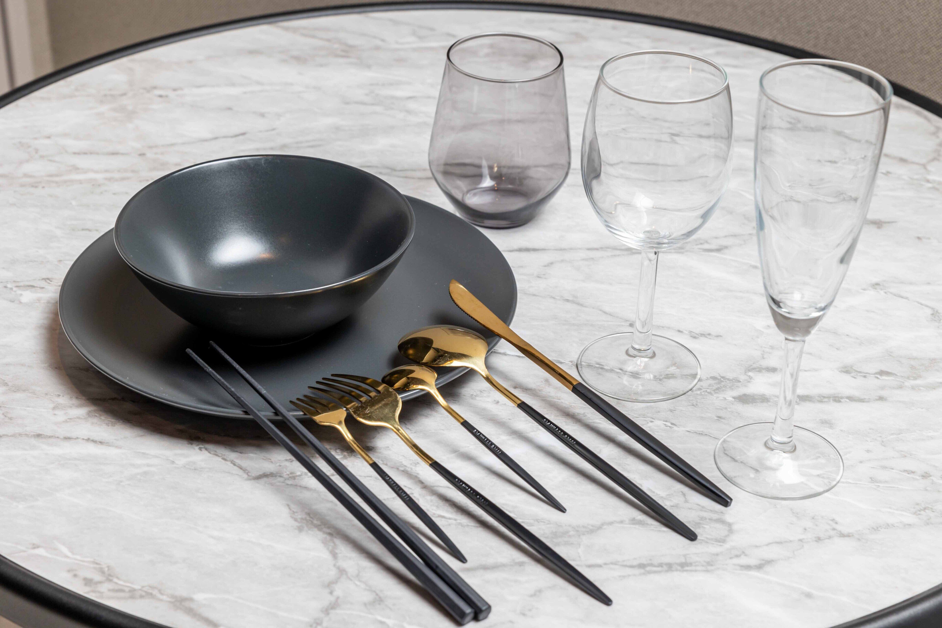 A table set with a black bowl, two wine glasses, and a set of cutlery including a gold spoon and black chopsticks.