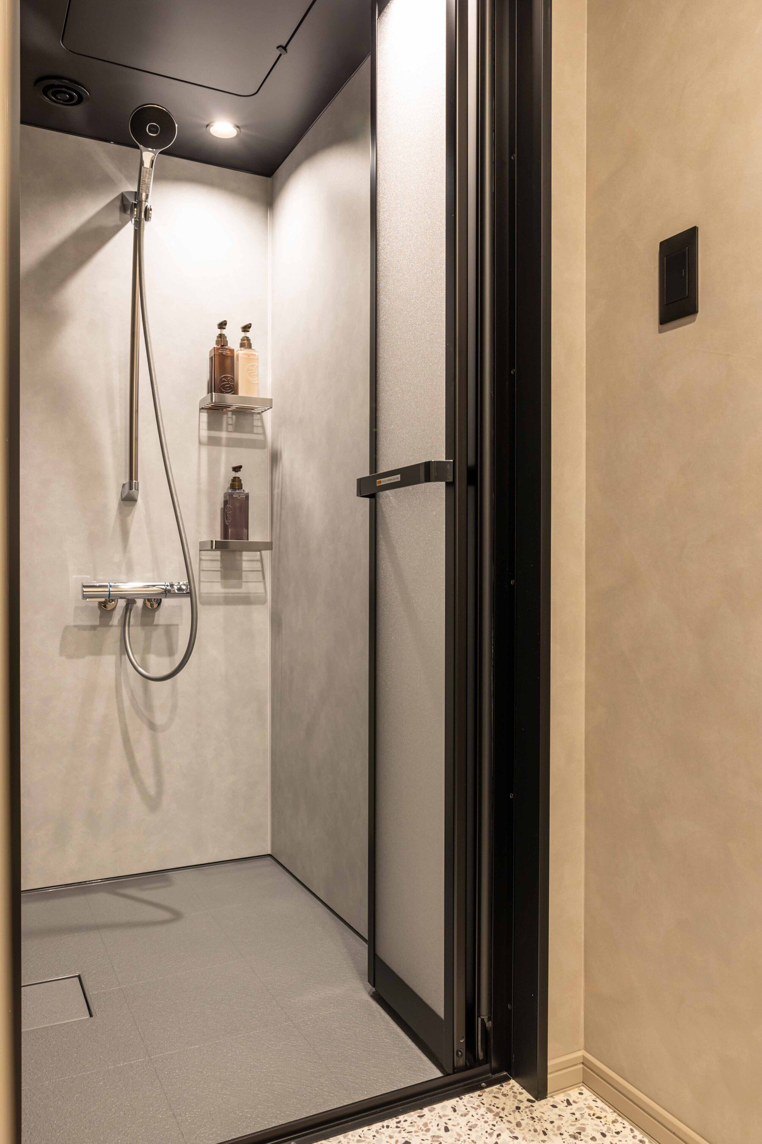 A walk-in shower with a glass door, a handheld showerhead, and a shelf with toiletries.
