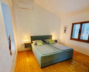 Ein Schlafzimmer mit einem Bett, Holzboden, zwei Nachttischen, einer Wand-Klimaanlage und einem Fenster mit Jalousien.