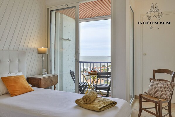 A hotel room with a bed, a nightstand, a lamp, a chair, and a balcony with a view of the sea.