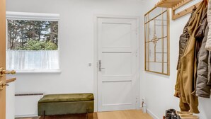 Une pièce avec une porte blanche, un banc, un porte-manteau avec des vestes, et une fenêtre donnant sur des arbres.