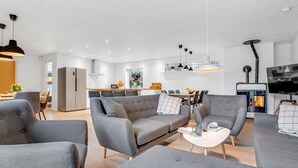 Un salon moderne avec des canapés gris, une table basse et un téléviseur. Au fond, on aperçoit une cuisine avec un coin repas.