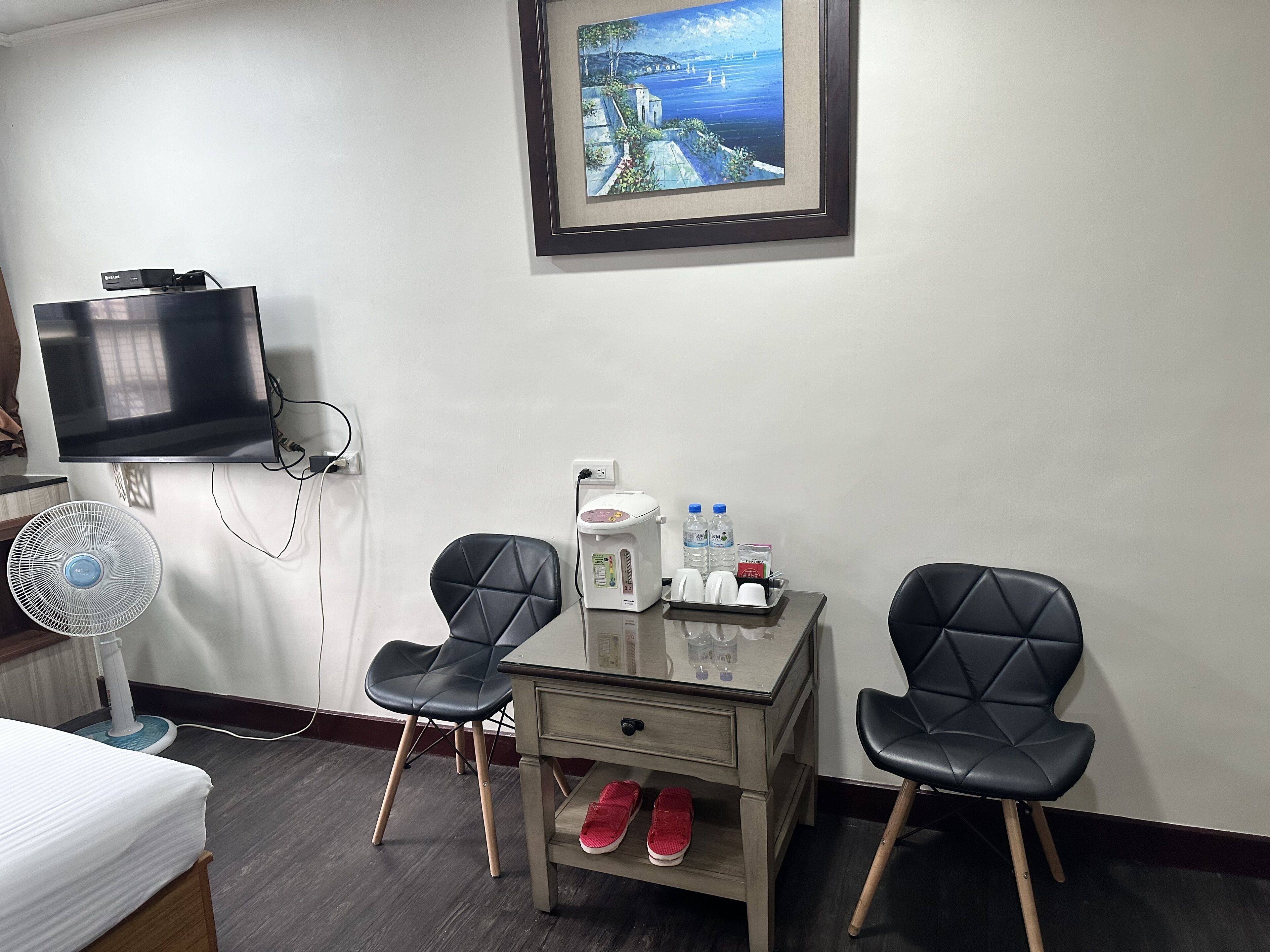 A hotel room with a bed, a desk with two chairs, a TV, a fan, a scale, and a framed picture on the wall.