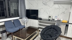 A modern living room with a glass-top coffee table, two blue armchairs, a black pouf, a standing fan, a flat-screen TV, a kitchen area with a marble backsplash, and a compact refrigerator. - JianGuo (Luodong)