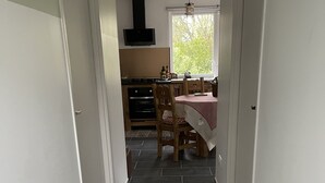 Een smalle gang met donkere betegelde vloer die leidt naar een keuken met houten kasten, een fornuis en een eettafel met stoelen.