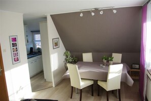 A dining area with a table and chairs, a vase with flowers, and a kitchen in the background.