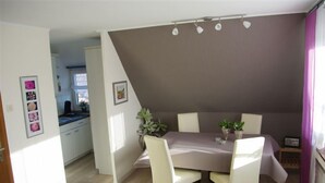 A dining area with a table set for four, surrounded by chairs. There is a small plant on the table, and the room has a modern design with a sloped ceiling and pendant lights. - Vacation apartment Wenge, 1-2 pers. (Ascheberg-Davensberg)