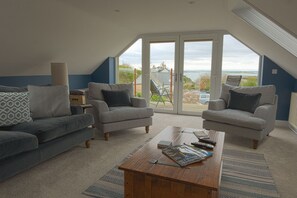 A living room with a high ceiling, two armchairs, a sofa, a coffee table with remote controls, and a view of the outdoors through large windows.