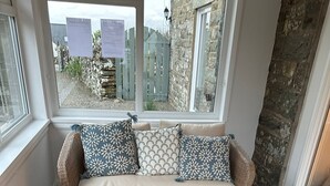 A room with a herringbone-patterned floor, a wicker sofa with patterned cushions, a small wicker basket, and a window with a view of a stone wall and a wooden fence. - Owl Cote -  a cottage that sleeps 5 guests  in 3 bedrooms (Gatehouse of Fleet)