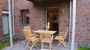A patio with a wooden table and chairs, a brick wall, and a window. - Ferienwohnungen Bisping33, Workershome 1-5 Pers (Ascheberg)
