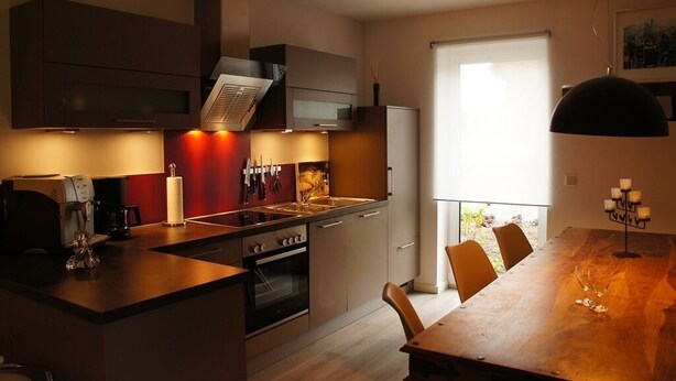A modern kitchen with a central island, built-in oven, and stainless steel appliances. There is a dining area with a large table and chairs, and a window providing natural light. - Ferienwohnungen Bisping33, Workershome 1-5 Pers (Ascheberg)