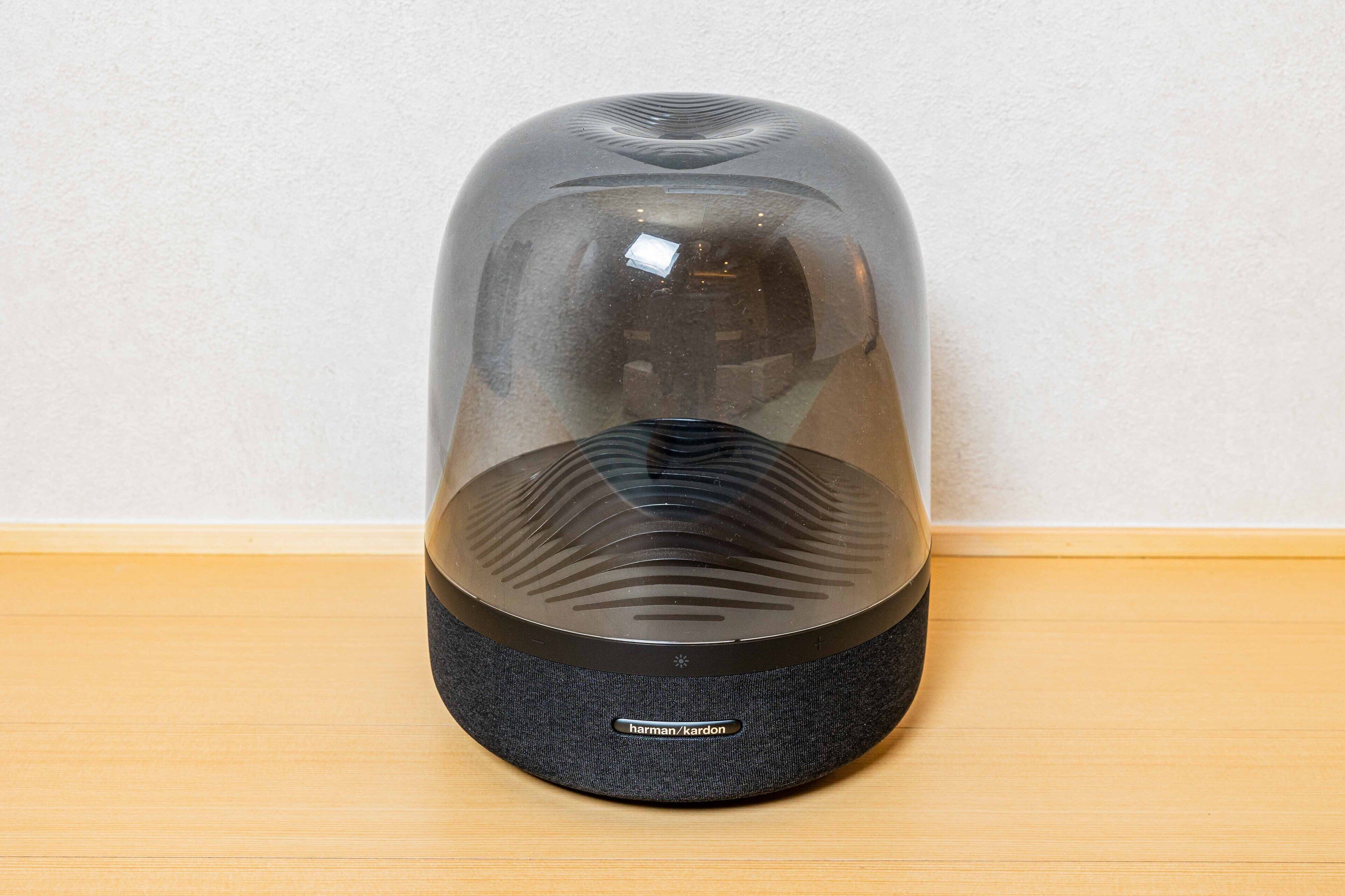 Un appareil noir, en forme de dôme, doté d’une base en tissu et d’un sommet transparent, portant l’inscription « harman/kardon », posé sur une surface en bois devant un mur uni.