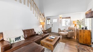 A spacious living room with a brown leather sofa, a wooden coffee table, and a beige armchair. A staircase is visible in the background.