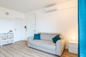 Een kamer met een beige bank, een witte nachtkastje, een wandairconditioner en een deur.