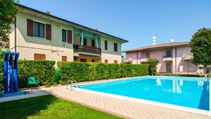 Une piscine avec une terrasse carrelée, une pelouse et un bâtiment doté de balcons.