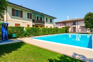 Une piscine avec une terrasse carrelée, une pelouse et un bâtiment doté de balcons.
