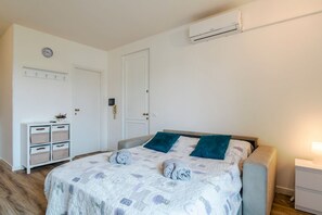 Une chambre à coucher comprenant un lit, une table de chevet, une commode, une horloge murale, une étagère avec des boîtes de rangement et un appareil de climatisation.