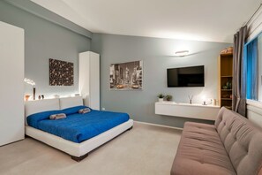 A modern bedroom with a bed, a sofa, a TV, and a small shelf with decorative items.