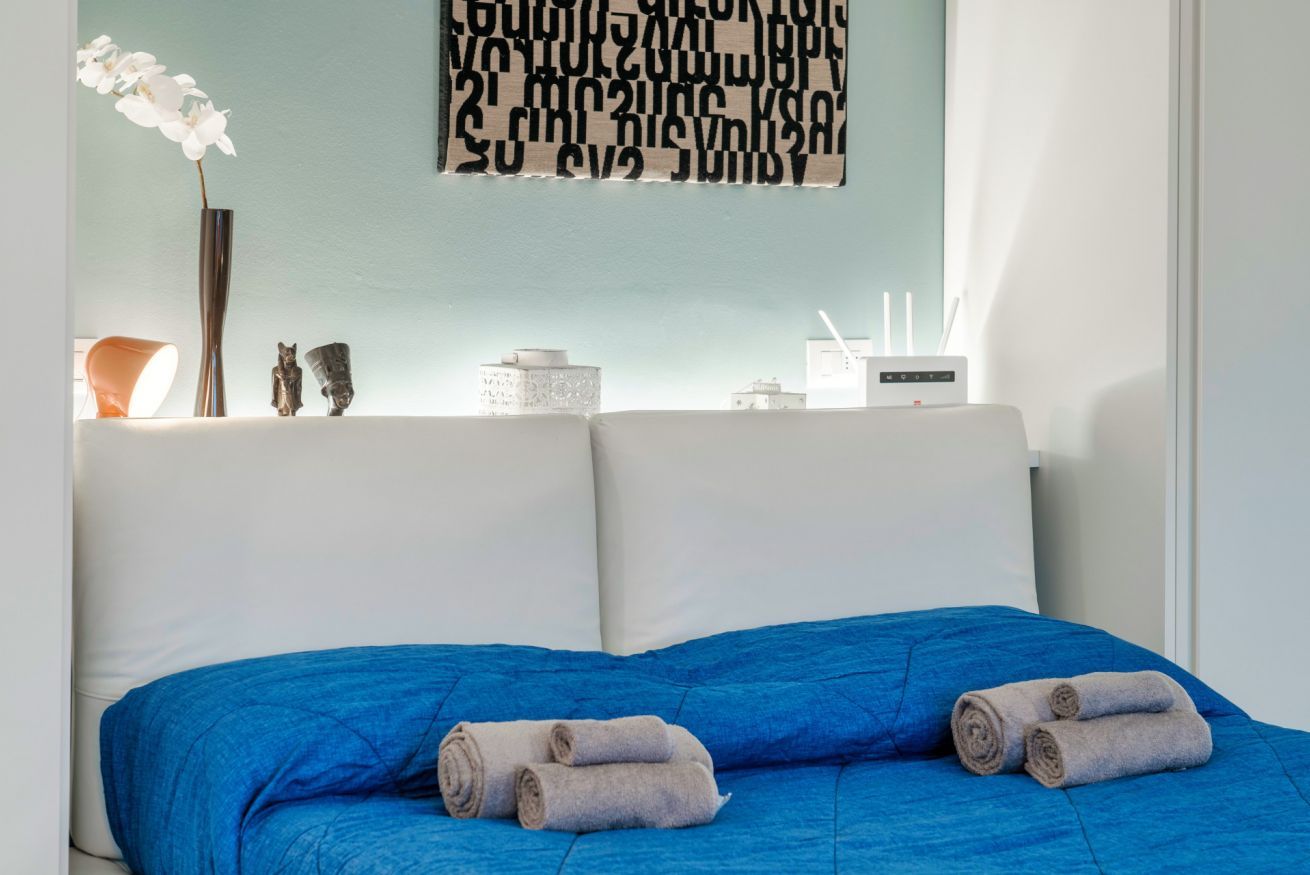 A neatly made bed with blue bedding and two rolled towels, a vase with white flowers, and a wall with a geometric pattern.