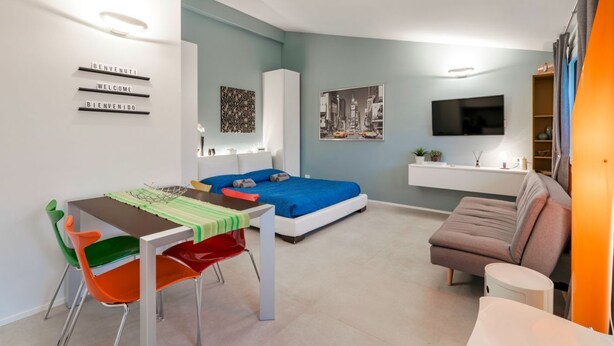 Un espacio habitable moderno y funcional con cama, mesa de comedor, sillas de colores, sofá, televisor y obras de arte en las paredes.