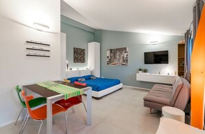 A modern, compact living space with a bed, a dining table, colorful chairs, a sofa, a TV, and artwork on the walls.