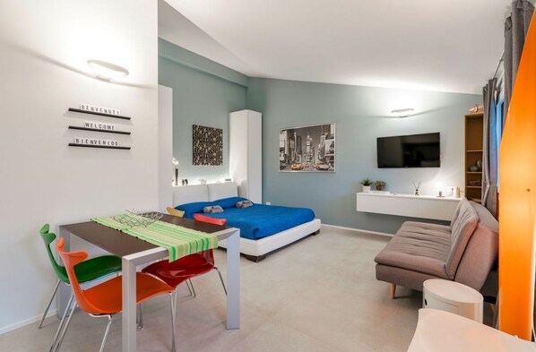 A modern, compact living space with a bed, a dining table, colorful chairs, a sofa, a TV, and artwork on the walls.
