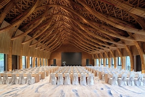 Un salón amplio con filas de mesas y sillas, un escenario y un techo alto de madera.