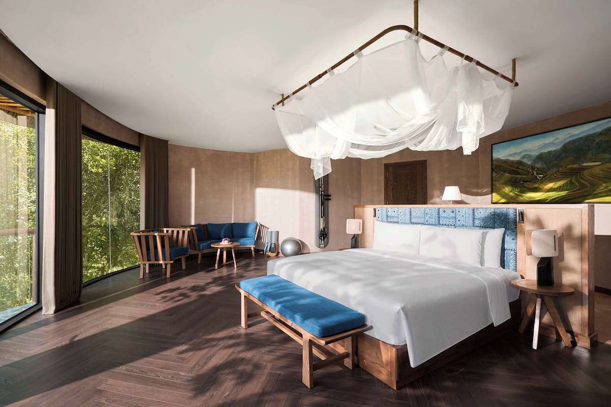 Un dormitorio moderno con una cama grande, un banco, una silla y un escritorio. Hay un ventanal y un cuadro en la pared.