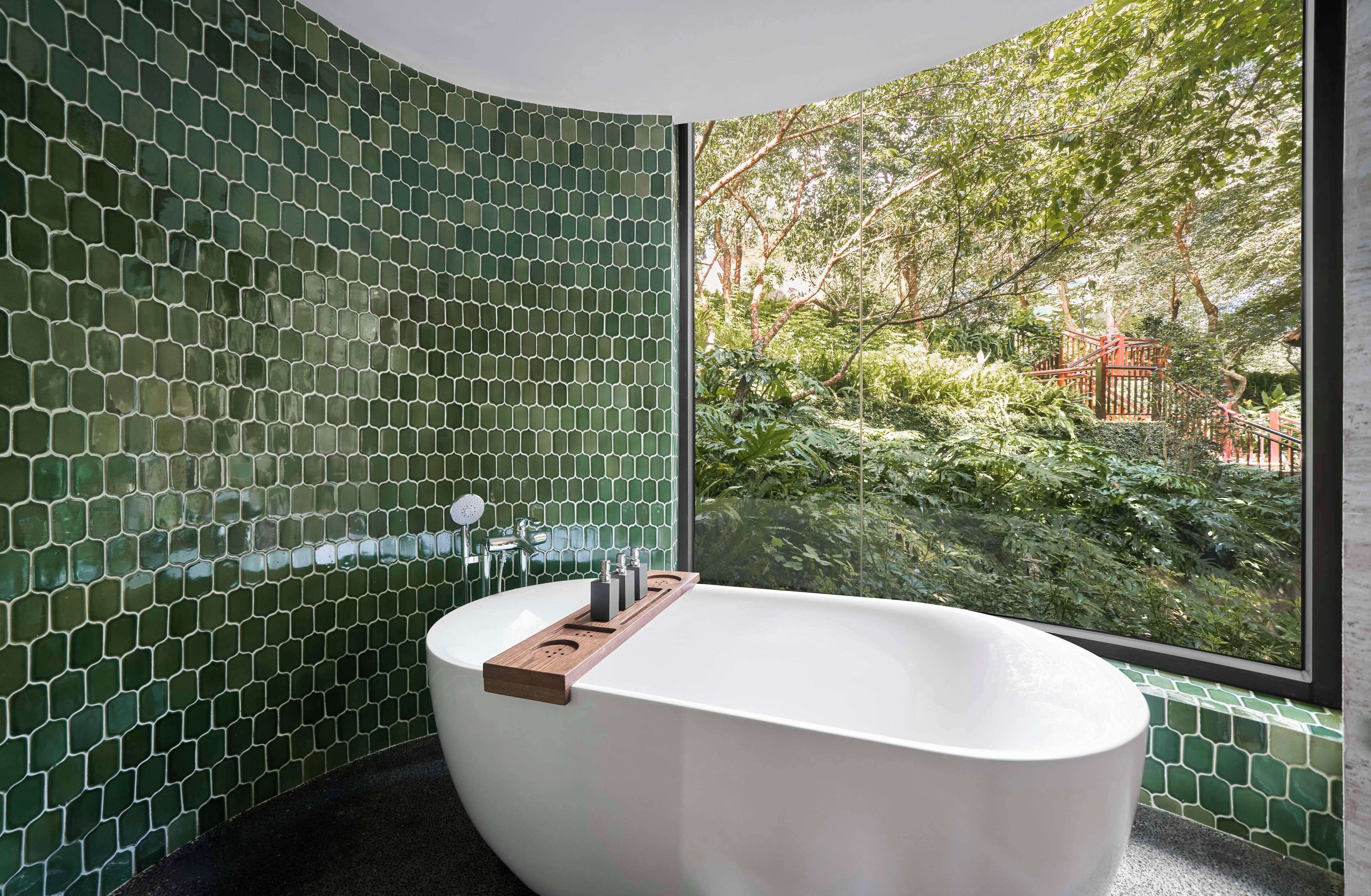 Um banheiro moderno com banheira livre, azulejos de mosaico verde e vista para um jardim exuberante através de uma grande janela.